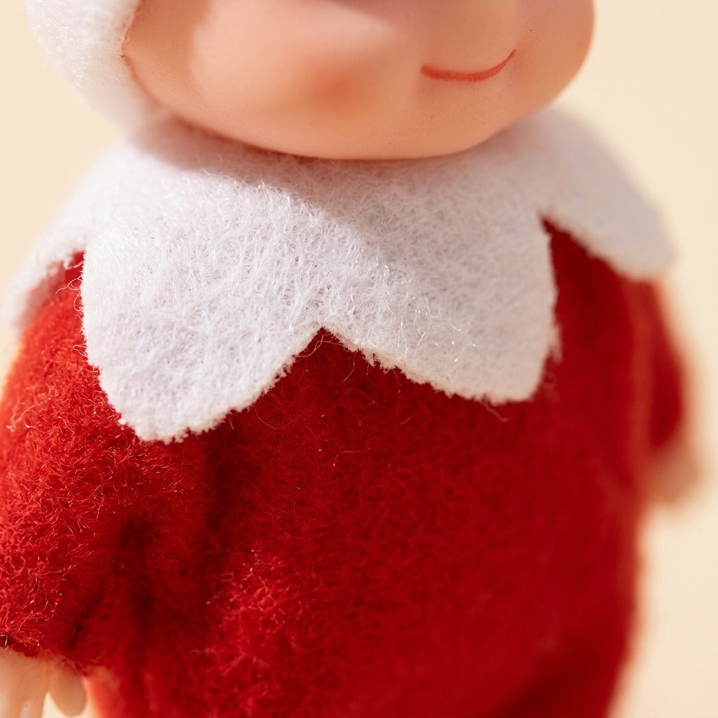 A close-up shows the elf doll’s textured red fabric outfit, scalloped white collar, smiling mouth, and small hand against a cream background.