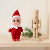 Elf doll in red clothing with white trim stands beside a wooden nutcracker and evergreen sprig on a pale wooden shelf.