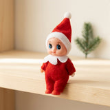 A red Christmas elf doll sits at the edge of a wooden shelf, with a blurred evergreen sprig behind it.