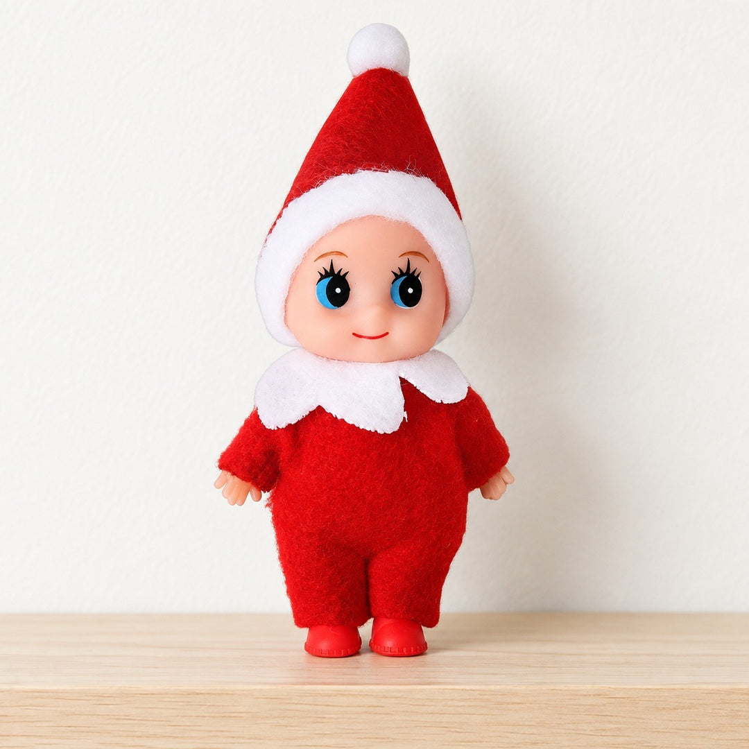 A blue-eyed Christmas elf doll in a red suit and white-trimmed pointed hat stands on pale wood against a white wall.