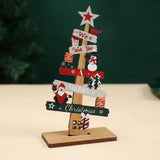 A wooden tabletop Christmas tree decoration with layered signs reading 'We Wish You a Merry Christmas,' decorated with red bird, snowman, Santa Claus, stars, and gift box illustrations, standing on a rectangular wooden base.