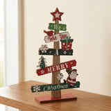 A wooden tabletop Christmas tree decoration with multiple painted sign-style branches reading phrases like 'We Wish You Merry Christmas' and decorated with a red star, bird, snowflakes, gift boxes, a small tree, and Santa Claus, shown on a wooden surface indoors.