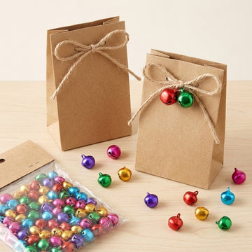 Brown paper gift bags with twine bows stand beside loose multicoloured jingle bells and a clear bag of bells on a wooden tabletop.