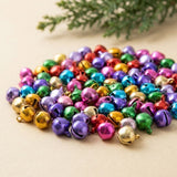 Shiny red, green, blue, purple, pink and gold jingle bells form a pile on a cream surface beneath evergreen branches.