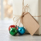 Red, green, and blue metallic jingle bells hang from twine beside a blank brown tag on a cream gift box.