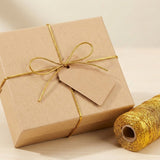 Metallic gold craft cord ties a kraft gift box and blank tag beside a matching spool on a light wood surface.