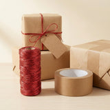 A red metallic craft cord spool stands beside brown packing tape and kraft paper parcels, including one tied with matching cord.