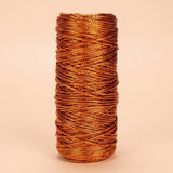 Copper-coloured craft cord with a metallic sheen is wound in overlapping strands on an upright spool against a pale peach background.