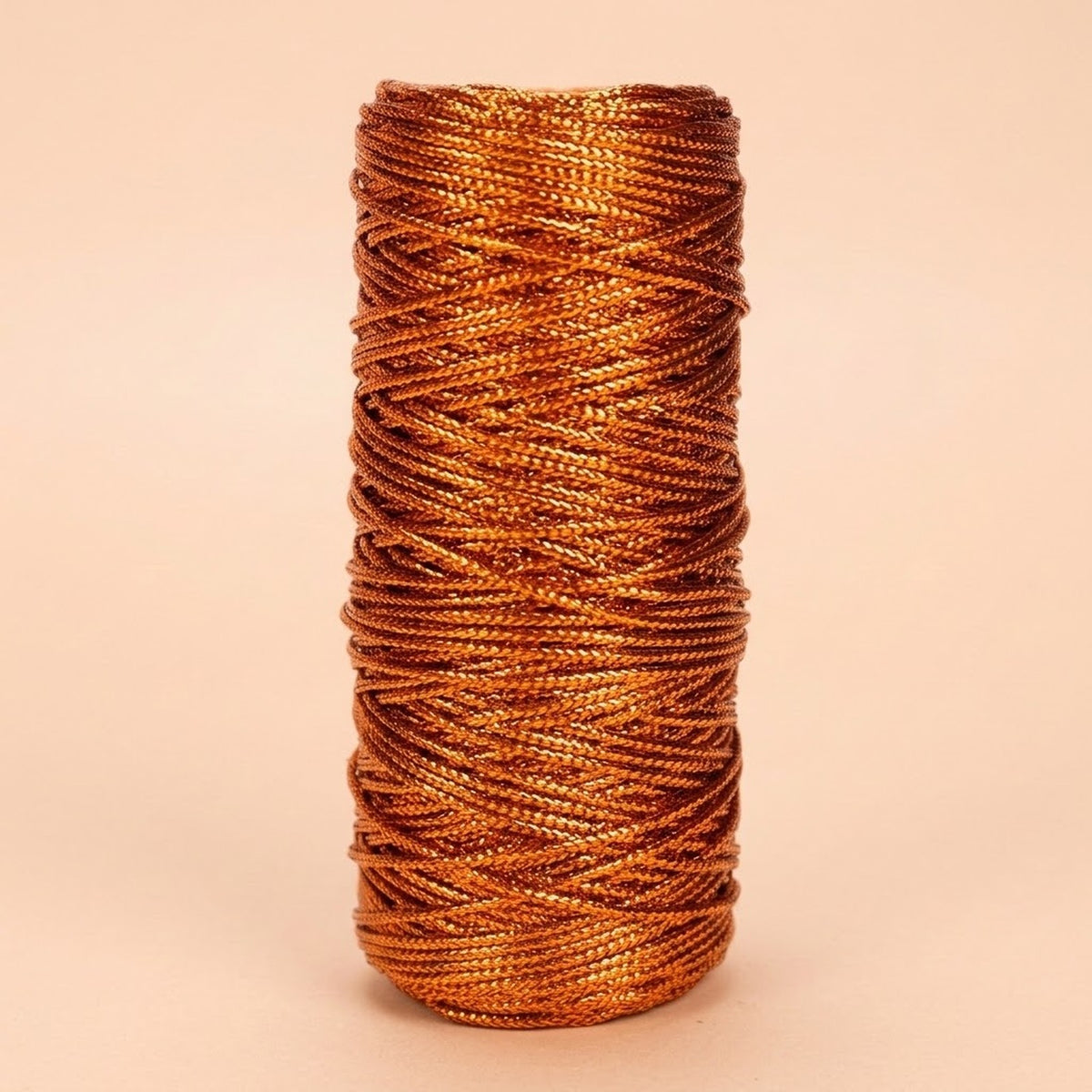 Copper-coloured craft cord with a metallic sheen is wound in overlapping strands on an upright spool against a pale peach background.
