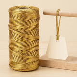 A gold metallic craft cord spool stands on pale wood beside a blank tag suspended from a wooden rod with matching cord.