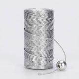 A silver metallic craft cord spool stands on a light grey background with its loose end threaded through a round silver bead.