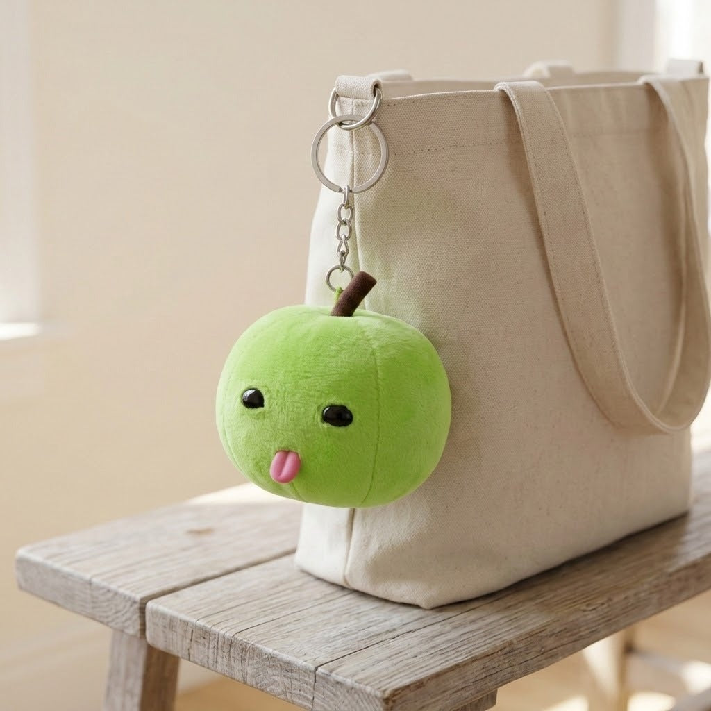 A green plush apple keychain with black eyes and a pink tongue hangs from a cream tote bag on a wooden stool.