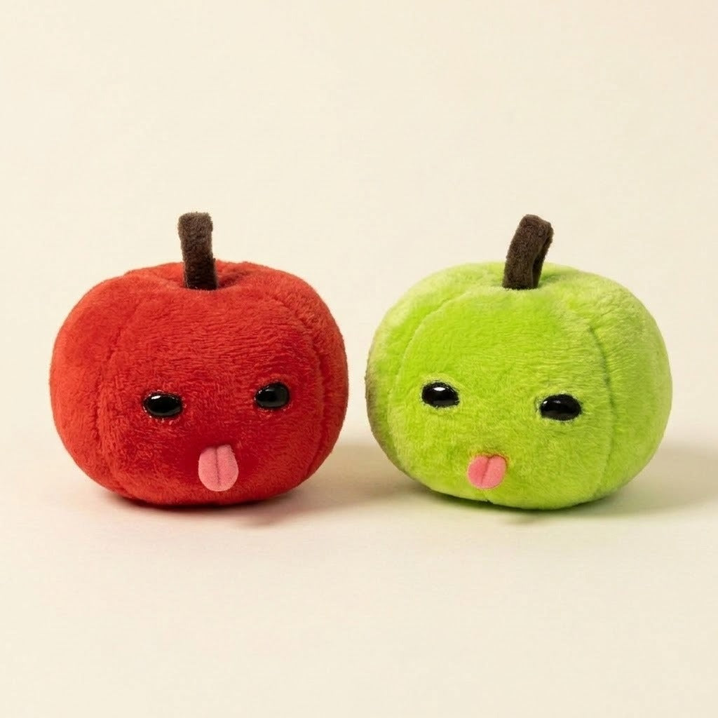 Red and green plush apple keychains with brown stems, black eyes, and pink tongues sit side by side against a cream background.
