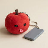 Red plush apple keychain with silver hardware sits beside a gray rectangular key fob on a cream surface.