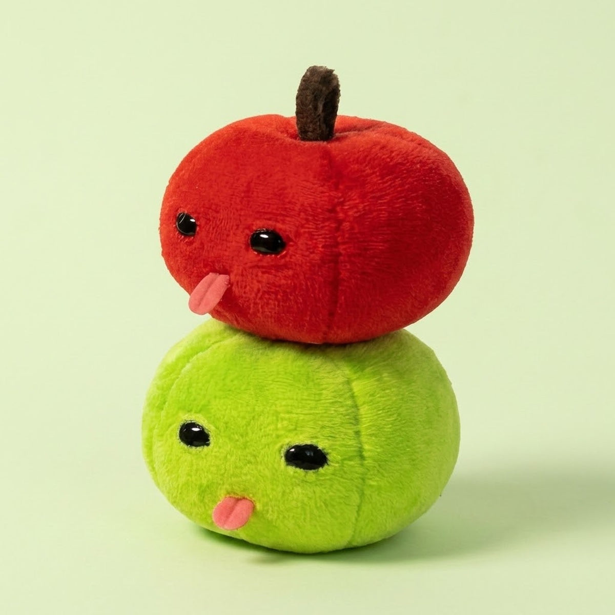 Red plush apple keychain rests atop a green plush apple keychain, both with black eyes and pink tongues, against a pale green background.