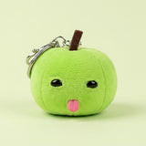 Green plush apple with a brown stem, black eyes, pink tongue, and silver keychain hardware sits against a pale green background.