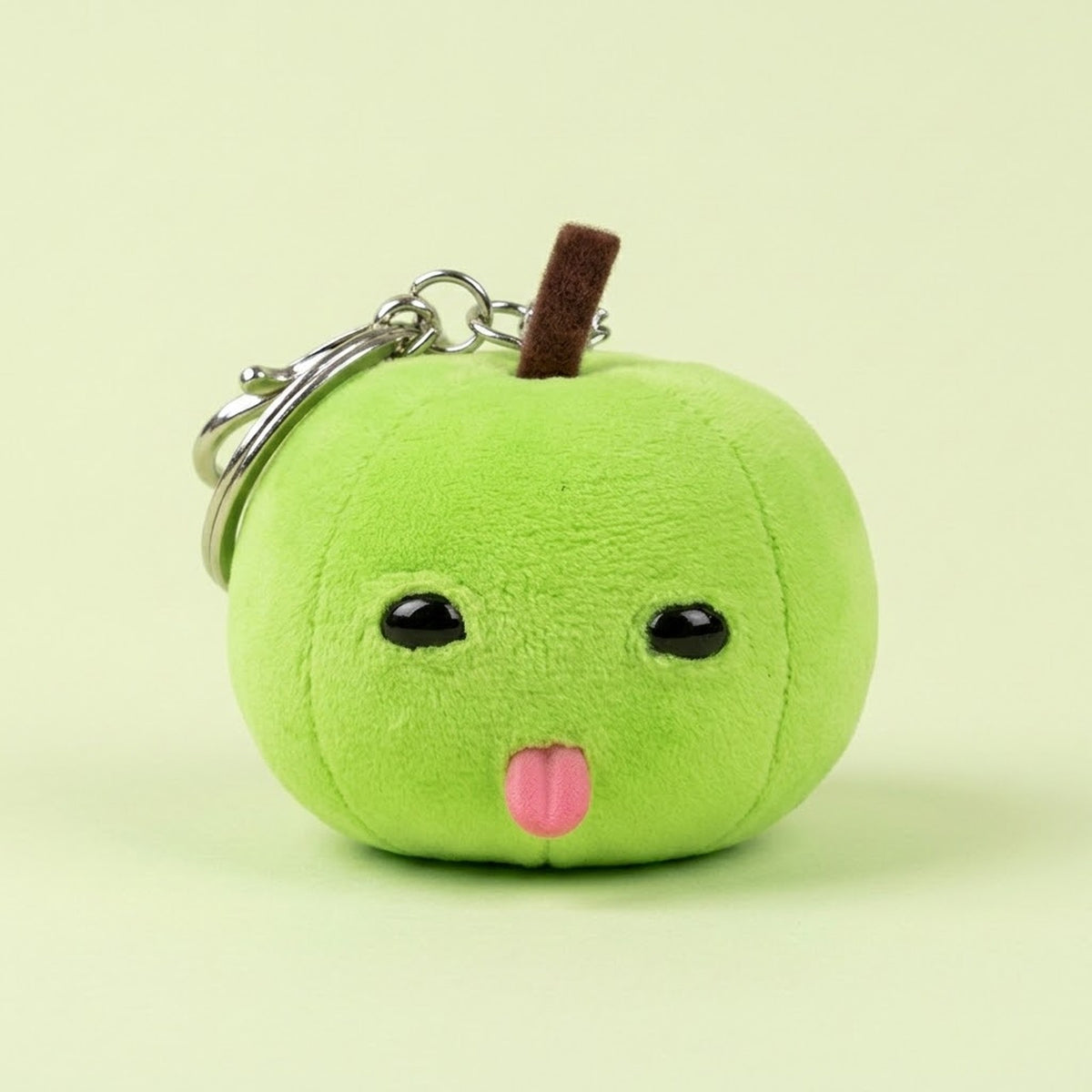 Green plush apple with a brown stem, black eyes, pink tongue, and silver keychain hardware sits against a pale green background.