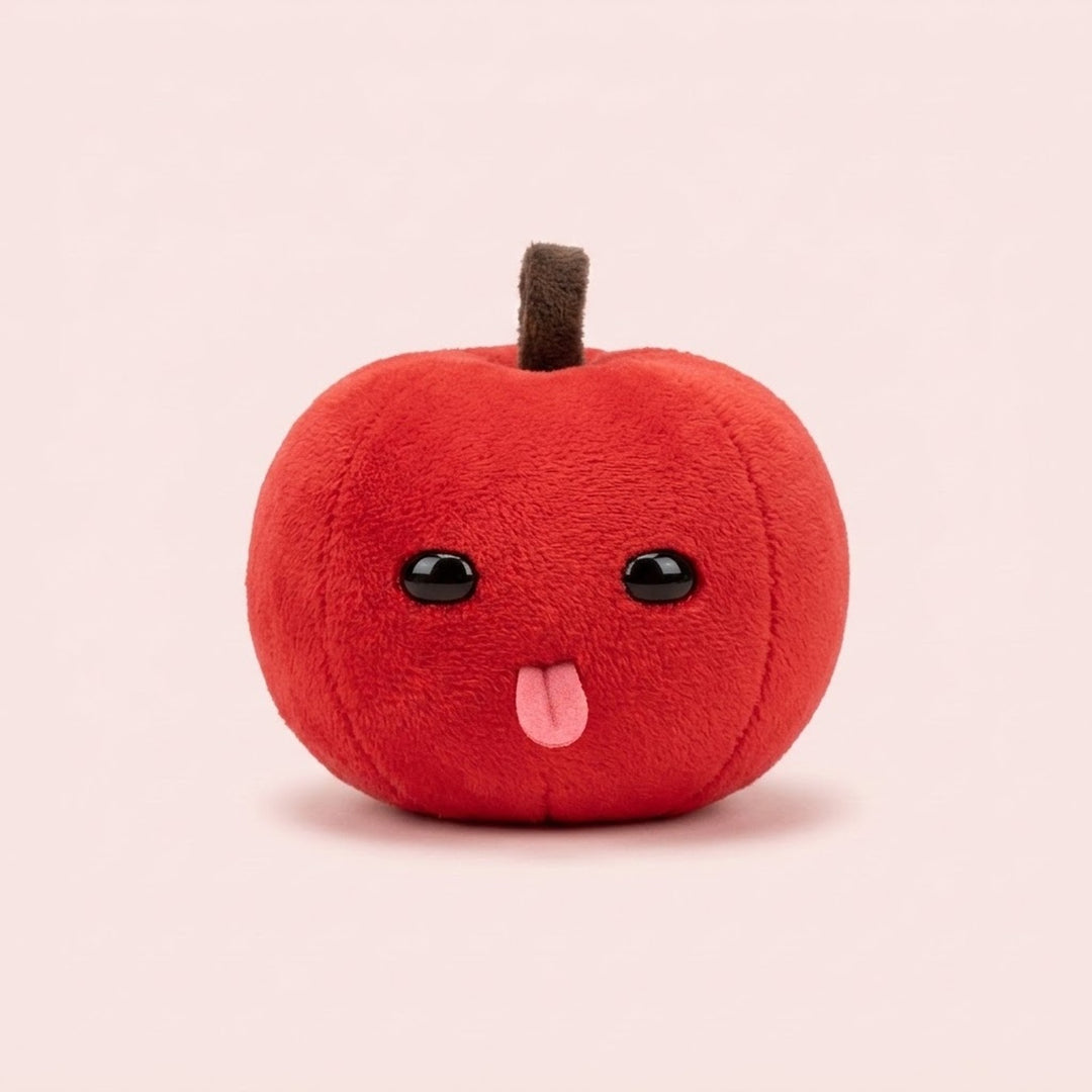 Red plush apple keychain with a brown stem, shiny black eyes, and pink tongue sits against a pale pink background.