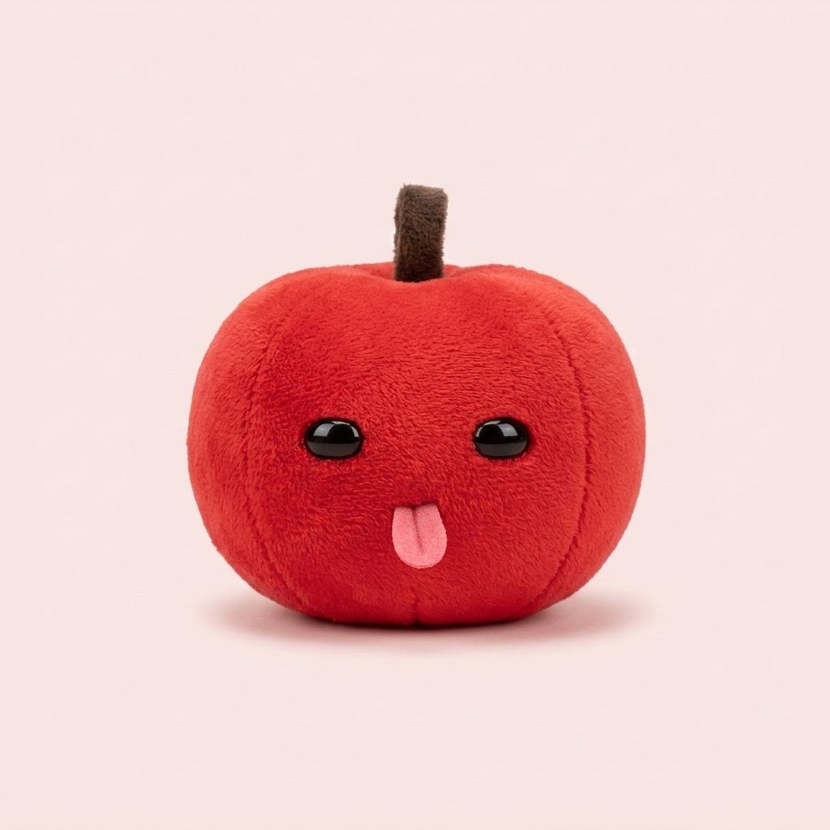 Red plush apple keychain with a brown stem, shiny black eyes, and pink tongue sits against a pale pink background.
