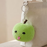 A green plush apple keychain hangs by a silver chain from a cream tote bag, showing black eyes and a pink tongue.