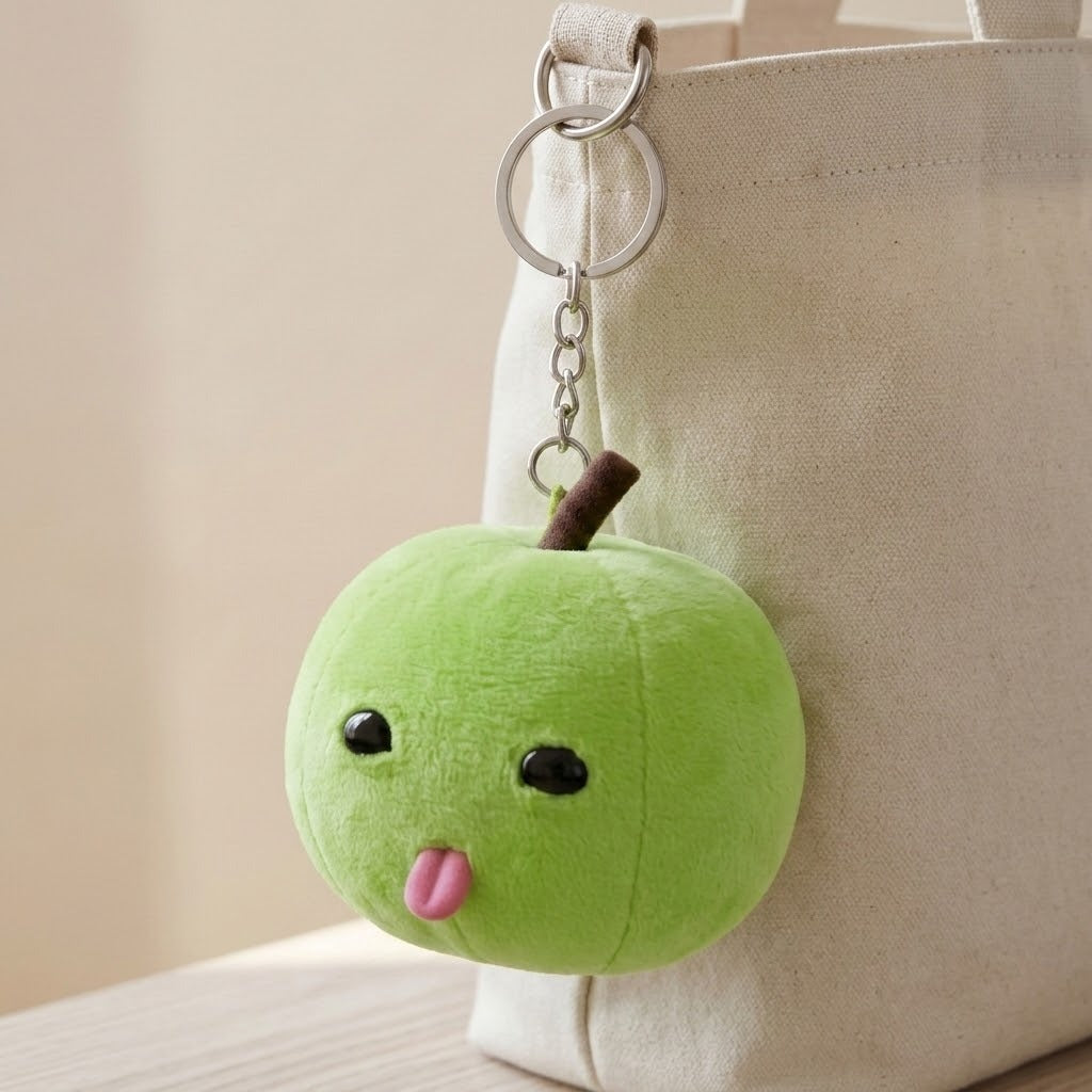 A green plush apple keychain hangs by a silver chain from a cream tote bag, showing black eyes and a pink tongue.