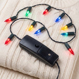 A light-up necklace with glowing multicoloured plastic bulbs, black twisted wire and a black battery box rests on cream cable-knit fabric.