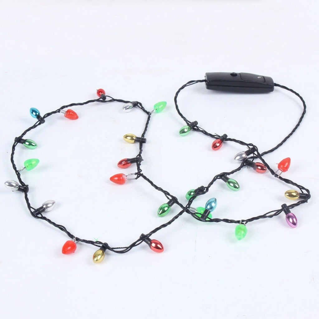 A light-up necklace with unlit multicoloured bulbs, black twisted wire and a rectangular black battery box lies loosely looped on white.
