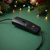 The light-up necklace’s black battery box has a round button and screw-fastened cover, resting on green beside blurred Christmas decorations.