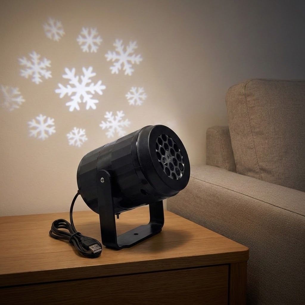 Black snowflake projector with USB cable sits on a wooden side table beside a beige sofa, casting white snowflakes onto the wall.