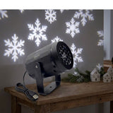 Black snowflake projector rests on a wooden console beside evergreen branches and miniature white houses, with white snowflakes projected behind it.