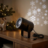 Black projector with coiled USB cable sits on a wooden cabinet beside reindeer ornaments and illuminated greenery, casting white snowflakes onto the wall.