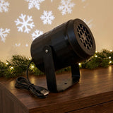 Black snowflake projector and coiled USB cable sit on a wooden cabinet beside illuminated evergreen garland, with white snowflakes on the wall.