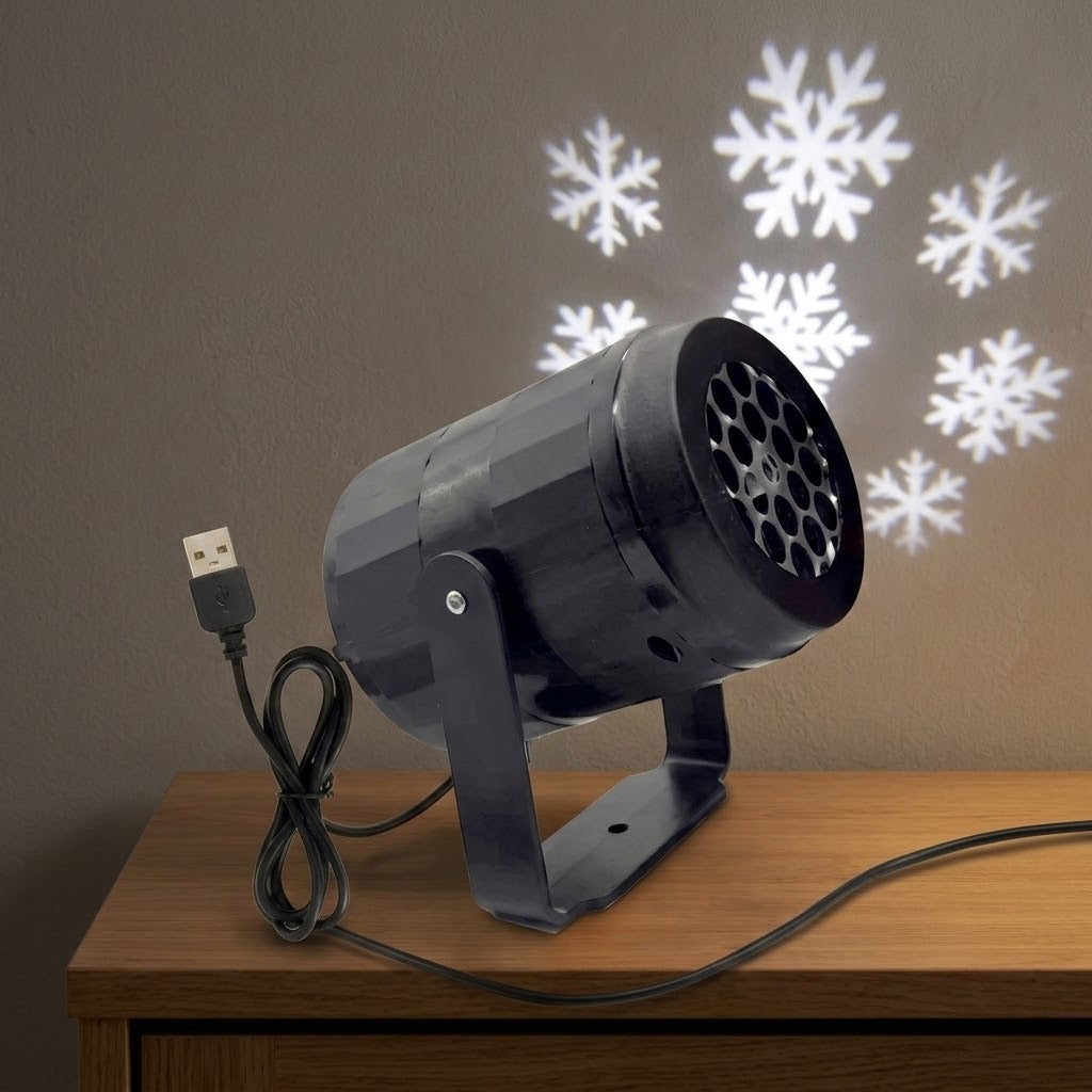 Black cylindrical projector with a USB cable and adjustable bracket sits on a wooden cabinet, with white snowflakes projected onto the wall behind.