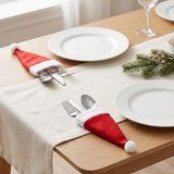 Two red Santa hat cutlery holders with white trim and pompoms hold silverware beside white plates on a wooden table with a cream runner.