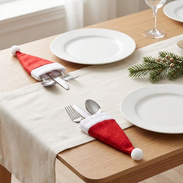 Two red Santa hat cutlery holders with white trim and pompoms hold silverware beside white plates on a wooden table with a cream runner.