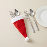A red cloth Santa hat cutlery holder with white trim holds a fork and spoon beside loose silverware and a white plate.