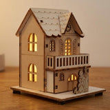 A light wooden Christmas house with illuminated arched windows, a balcony, and a roof clock stands on a wooden tabletop.