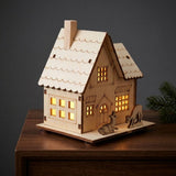 A glowing light wooden Christmas house with a chimney and deer silhouettes sits on a dark wooden cabinet against a charcoal wall.
