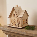 A pale wooden Christmas house with two deer figures stands on a rustic mantel beside an evergreen sprig.