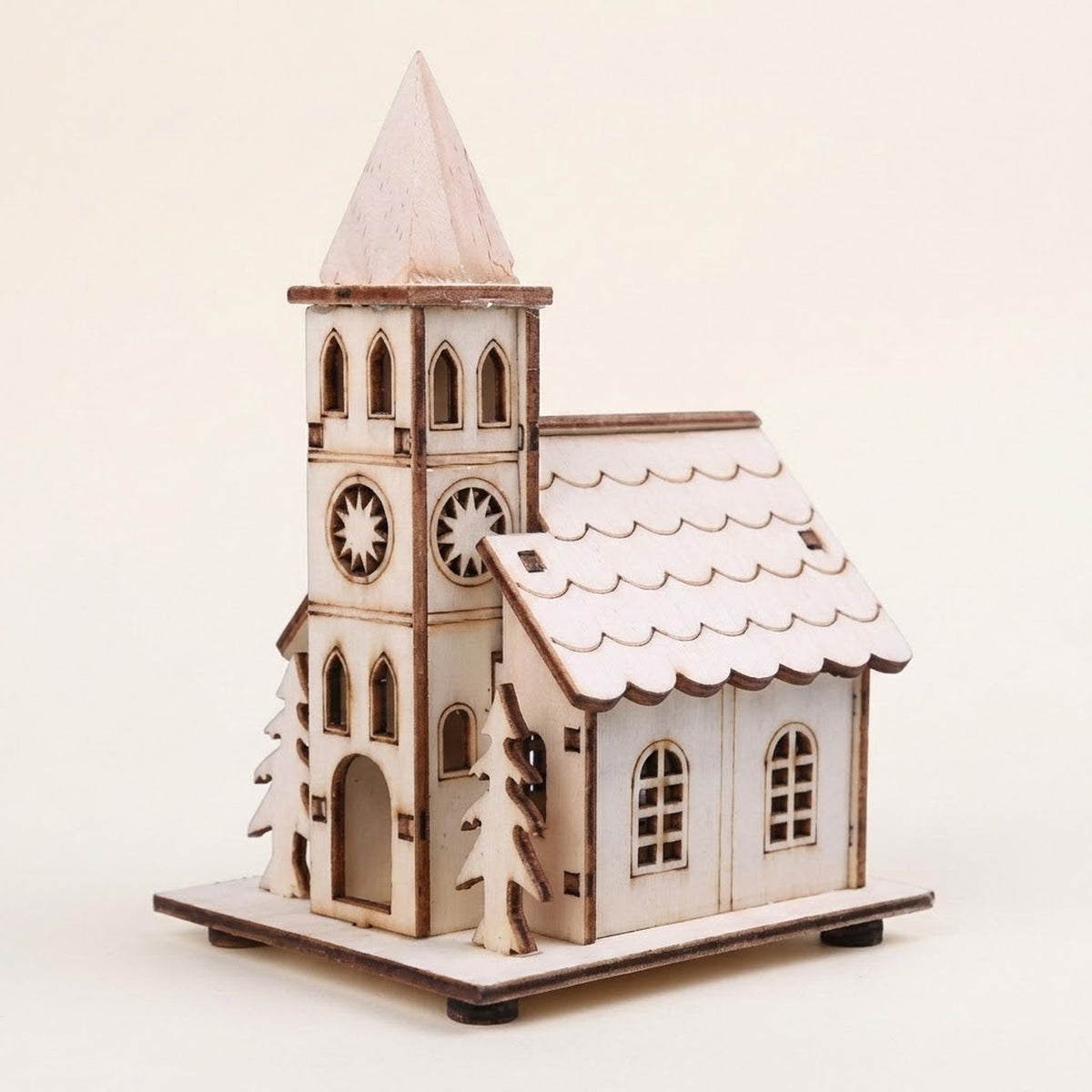 A pale wooden Christmas house with a pointed tower, starburst window cutouts, and fir trees stands against a cream background.