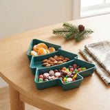 A dark green Christmas tree serving tray holds mandarin segments, almonds, and candies on a wooden table beside folded napkins.