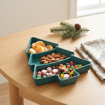 A dark green Christmas tree serving tray holds mandarin segments, almonds, and candies on a wooden table beside folded napkins.