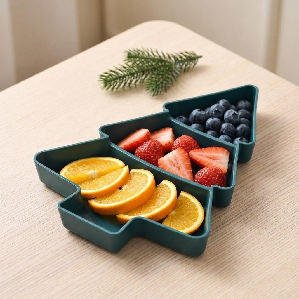 A dark green Christmas tree serving tray holds orange slices, strawberries, and blueberries in three compartments on a wooden table.