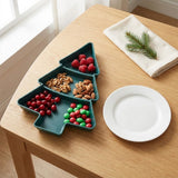 A dark green Christmas tree serving tray holds berries, nuts, and candies on a wooden table beside a white plate and folded napkin.