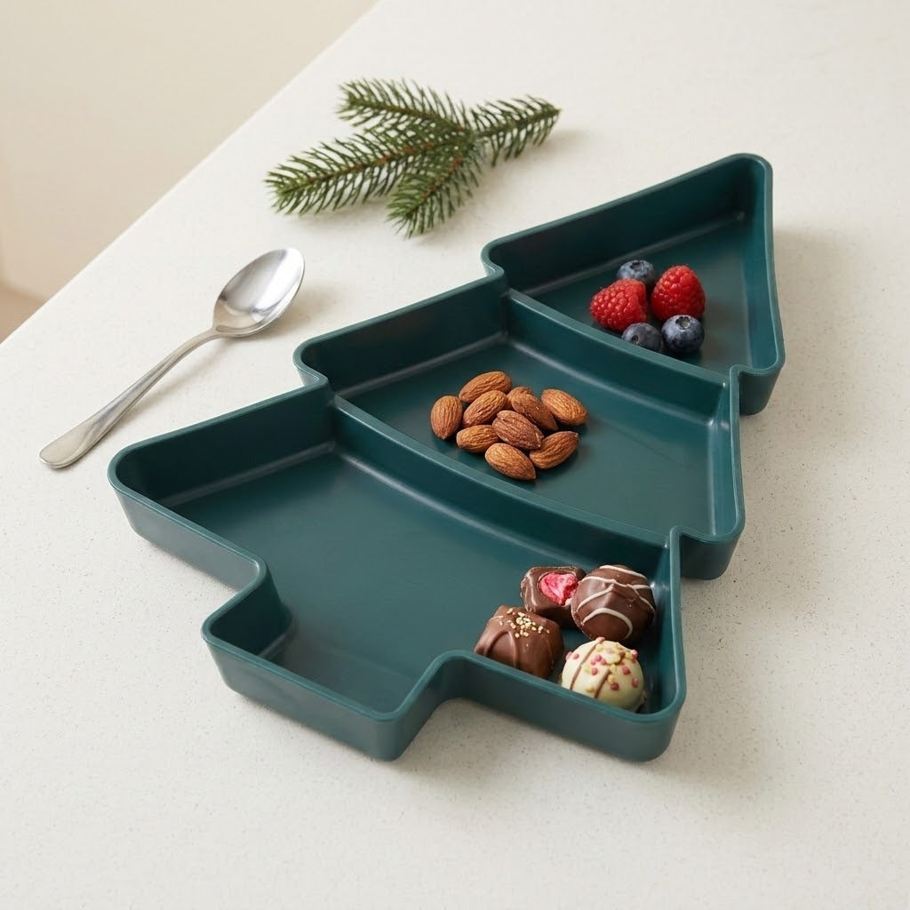 Dark green Christmas tree serving tray holds berries, almonds, and chocolates in three compartments on a pale tabletop beside a spoon and evergreen sprig.