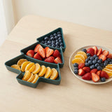 Dark green Christmas tree serving tray separates blueberries, strawberries, and orange segments into three compartments beside a round fruit plate on a wooden tabletop.