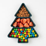 A dark green Christmas tree serving tray holds almonds, sliced strawberries, and colorful candies in three compartments against a white background.