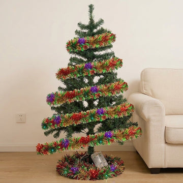 A red, green, and gold tinsel lighted garland spirals around a Christmas tree beside a cream armchair, with its clear battery box below.