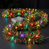 A red, green, and gold tinsel lighted garland glows with multicolored bulbs on a dark tabletop beside a clear battery box.