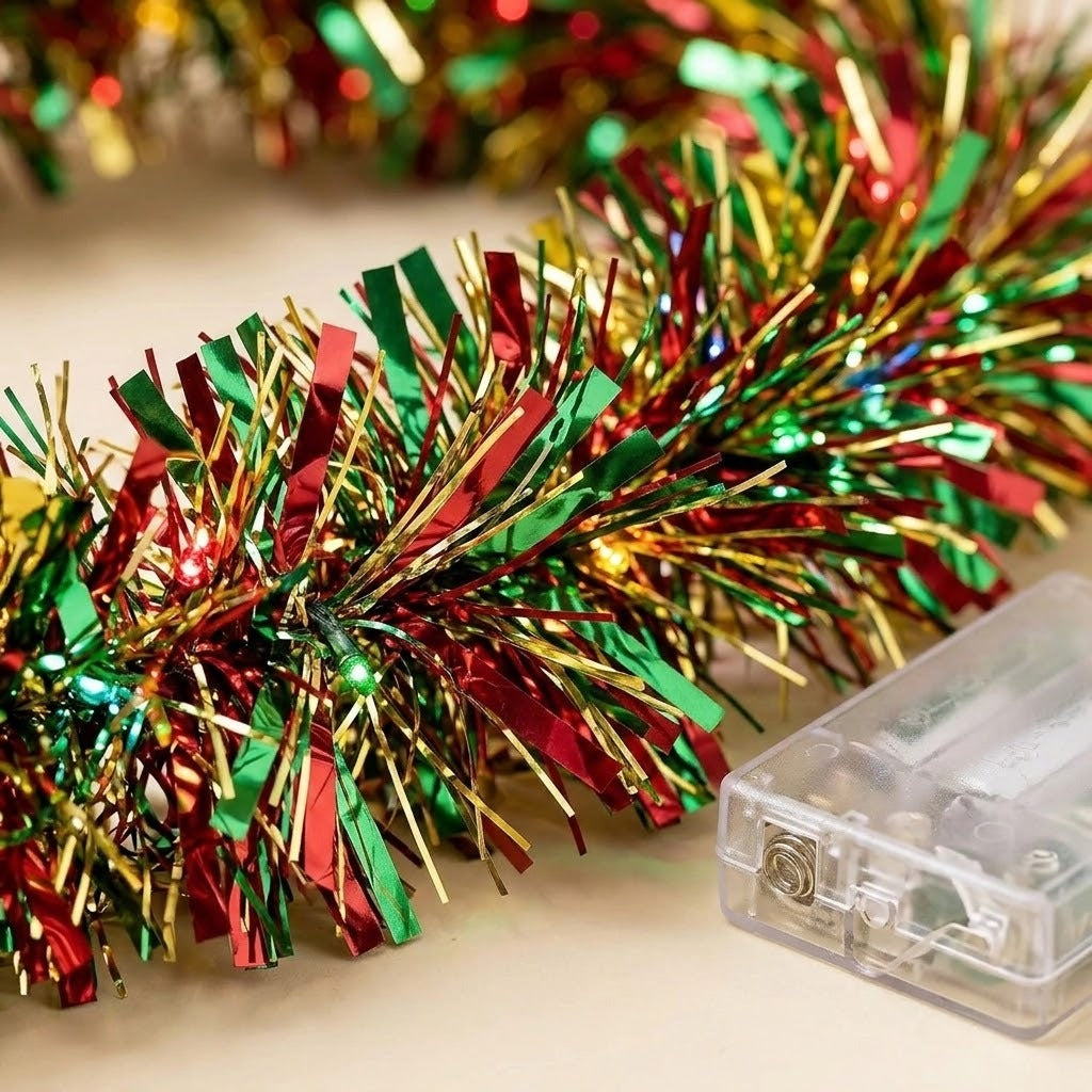 A close view shows shiny red, green, and gold tinsel on a lighted garland beside a clear battery box on a cream surface.